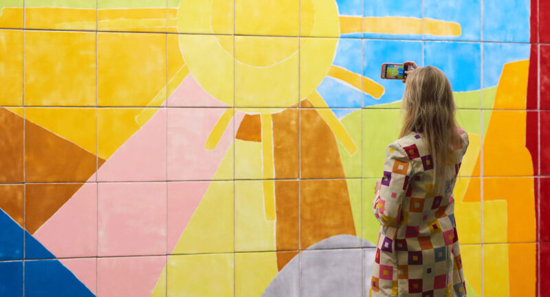 A woman photographs a colorful wall painting, shown from behind.